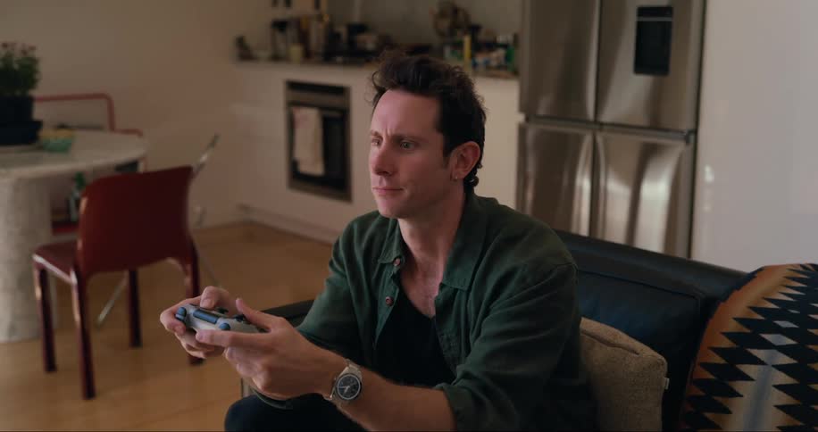 A man sitting on a black couch in a modern living space intensely focused while playing a video game with a controller with a kitchen and dining area visible in the background