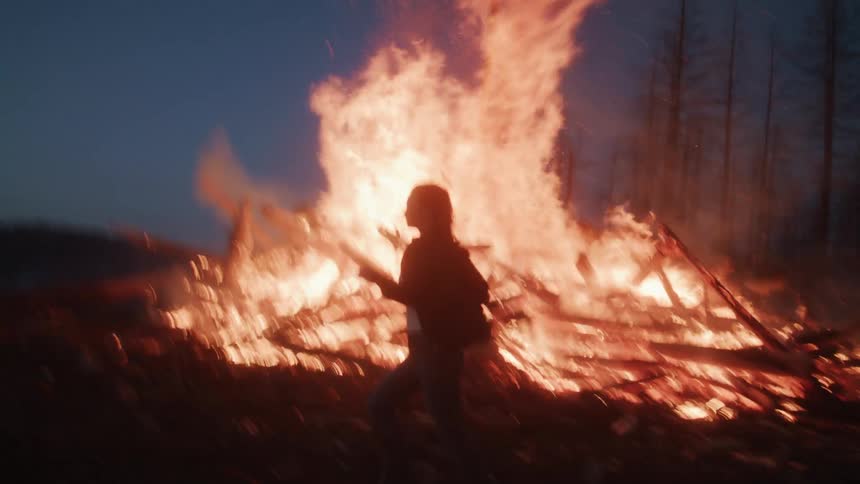 Woman running past a large bonfire in the forest at night