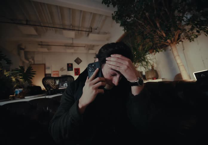 A person is sitting on a dark brown leather couch in a dimly lit room filled with various plants and decorations talking on a smartphone