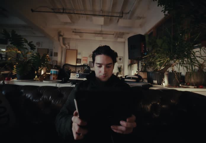 A person is sitting on a dark leather couch in a room filled with various plants books and electronic equipment holding a tablet