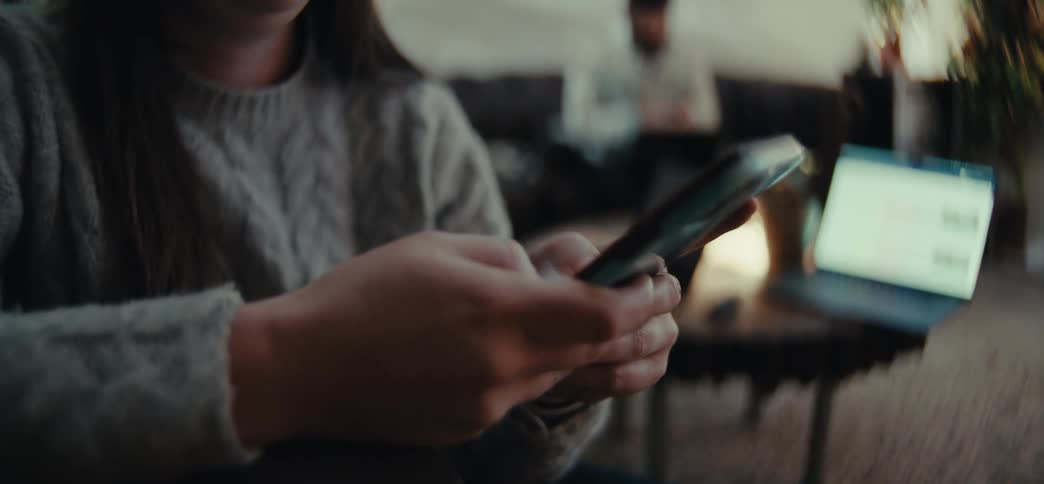 A close up of a woman texting on a smartphone with a laptop and a man sitting on a couch in the background