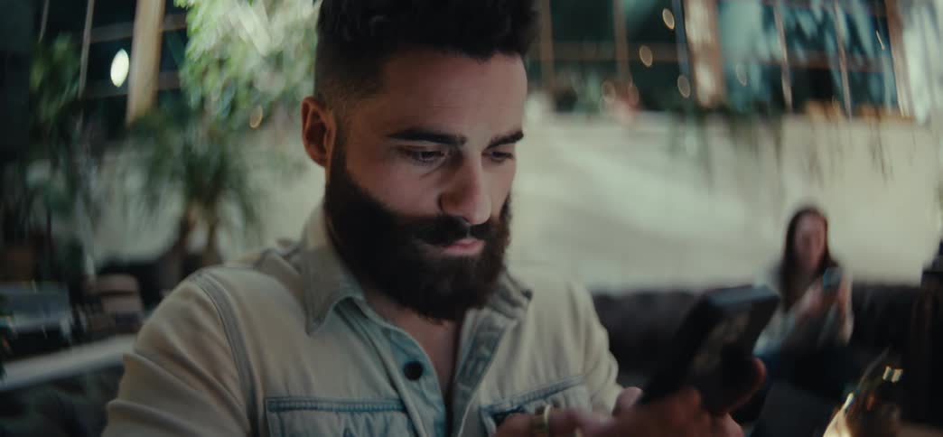 A bearded man is texting on a smartphone in an indoor setting with plants and lights in the background