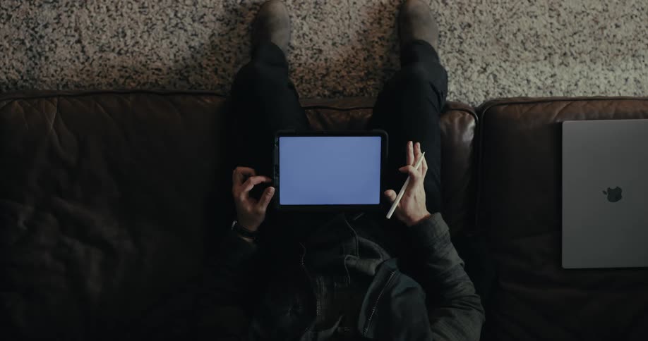 A person is sitting on a couch holding a tablet with a blank screen in one hand and a stylus in the other with a laptop placed beside them