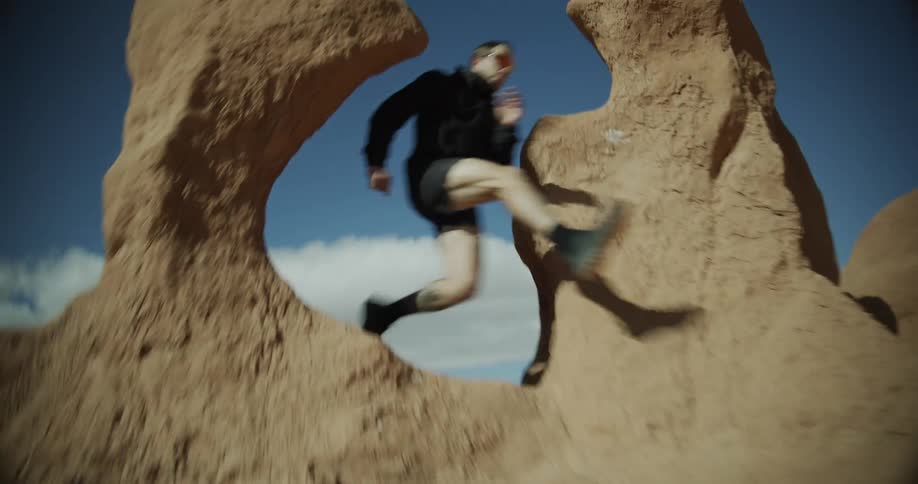 Male runner jumps through a gap in a rock formation in arid terrain on a cloudy summer day