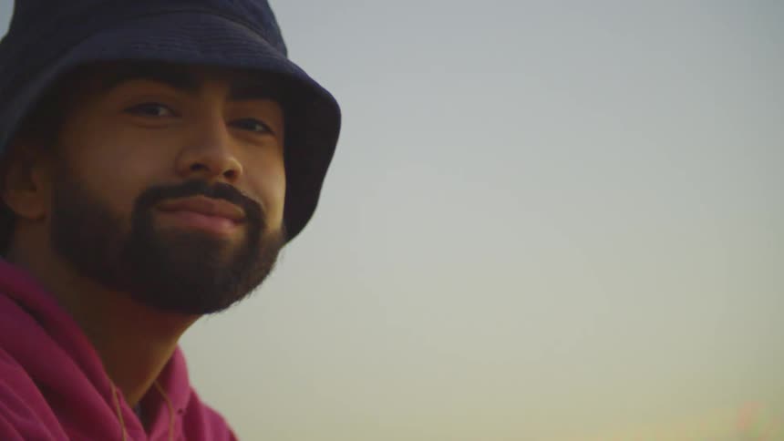 A person wearing a pink hoodie and a dark hat is sitting by the waterfront at dusk with palm trees on the left and a brightly lit ship in the background