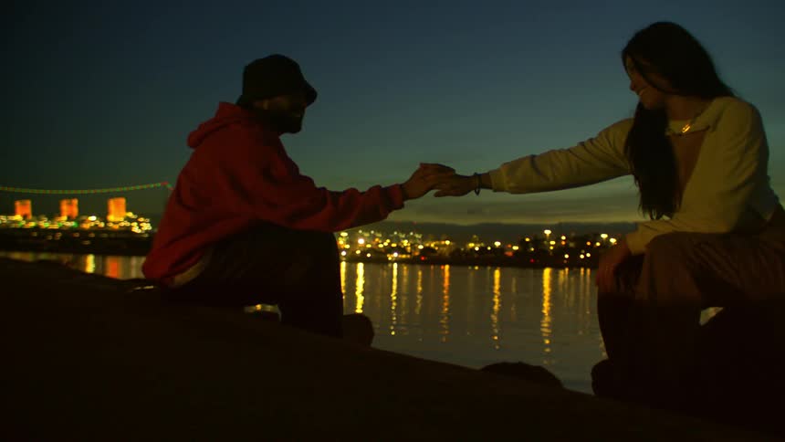 Man and woman sitting on the riverbank across from the brightly lit city at night reach out to touch hands