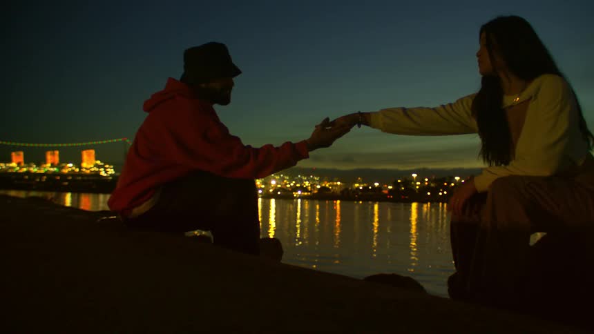 Man and woman sitting by the water at dusk reaching out their hands to each other with city lights reflecting on the river in the background