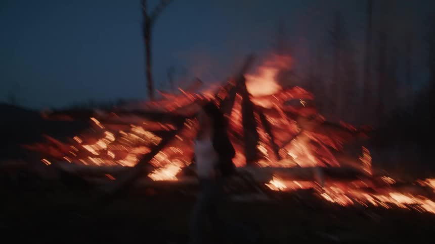 A woman runs next to a large bonfire with numerous logs burning intensely emitting bright flames and smoke against a dark nighttime background