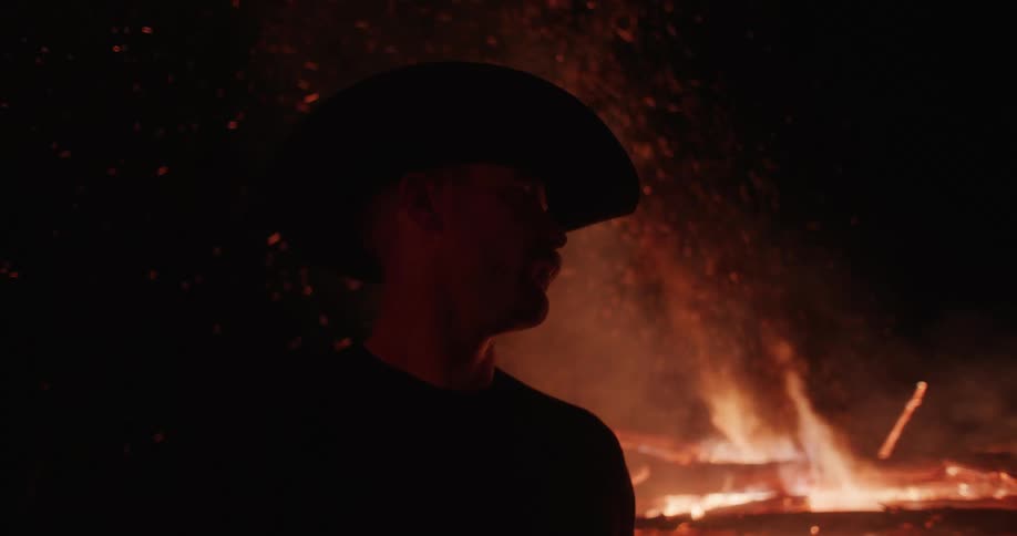 Silhouette of a man in a cowboy hat next to a roaring campfire outdoors at night