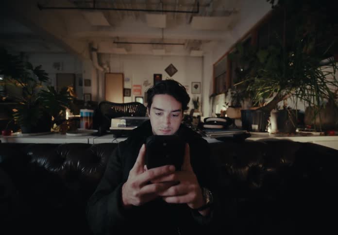 A person is sitting on a dark brown leather couch in a dimly lit room filled with various plants and decorations texting on a smartphone with both hands