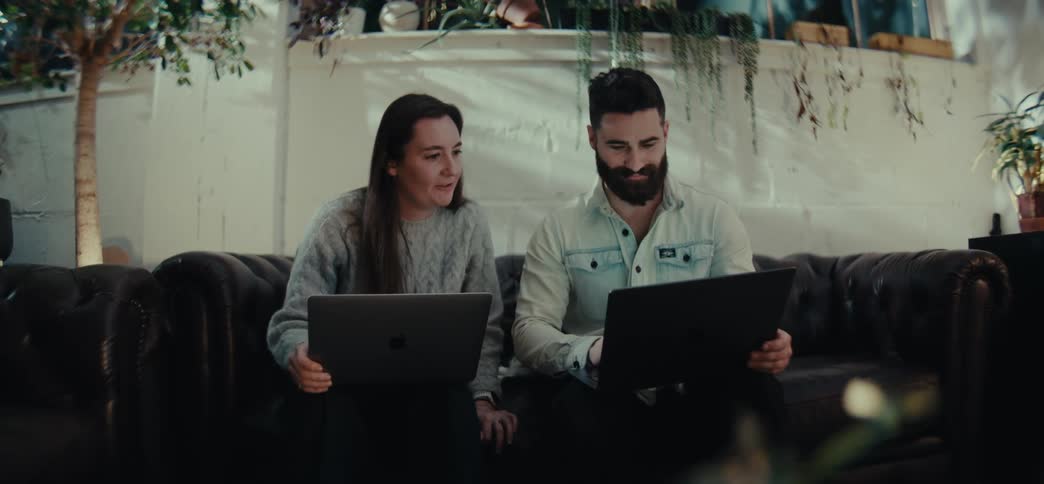 Man and a woman sitting in an armchair each with an open laptop and talking
