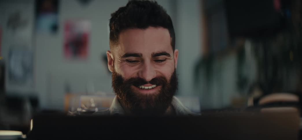 A young bearded man smiles and talks while typing on a laptop in an indoor setting with plants and lights around him