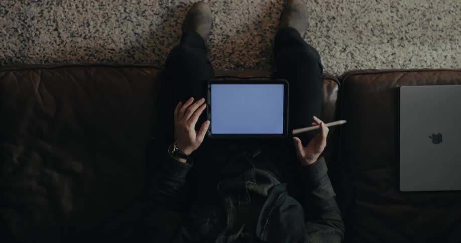 Person holding a tablet with a stylus pen sitting on a leather sofa with a laptop next to it
