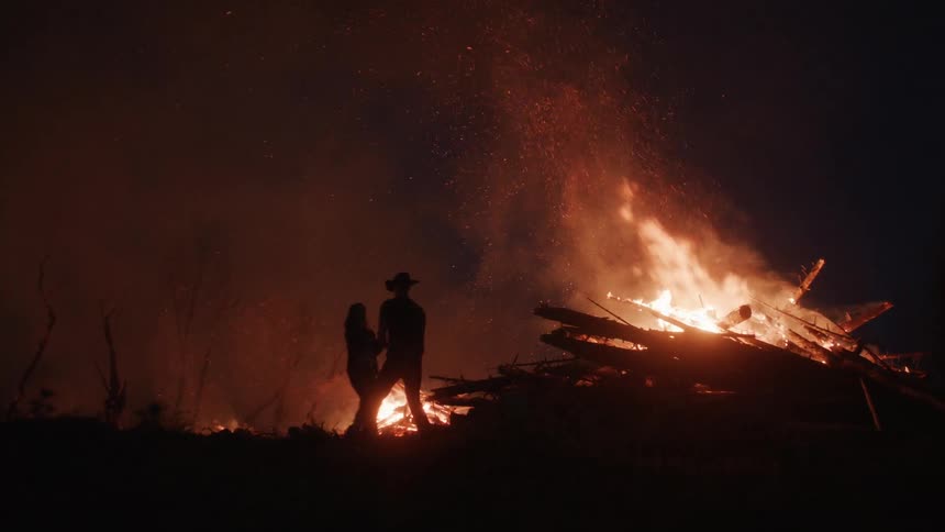 A couple dancing next to a large bonfire with numerous logs burning intensely emitting bright flames and smoke against a dark nighttime background
