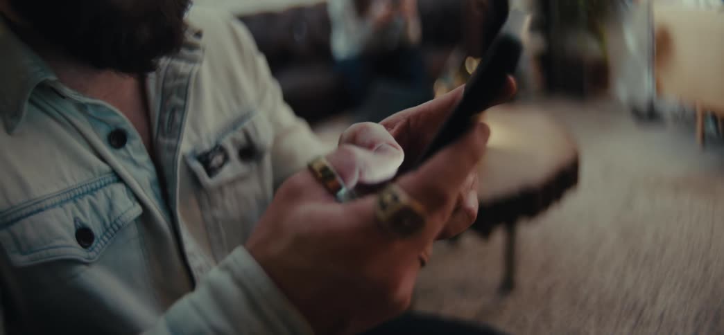 A bearded man with a light blue shirt uses a smartphone in the living room