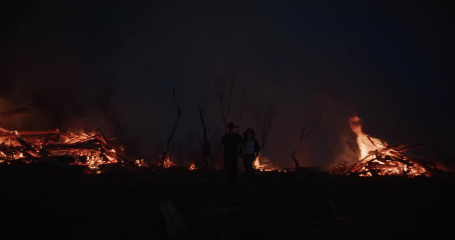 A couple runs away from several large bonfires with numerous logs burning intensely emitting bright flames and smoke against a dark nighttime background