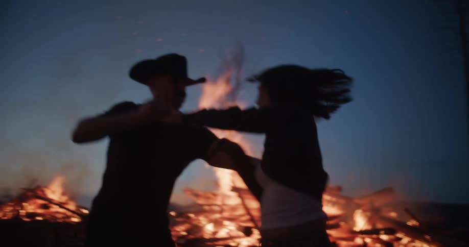 A silhouetted couple running together and sharing a passionate kiss framed against the dramatic glow of a roaring fire under the evening sky