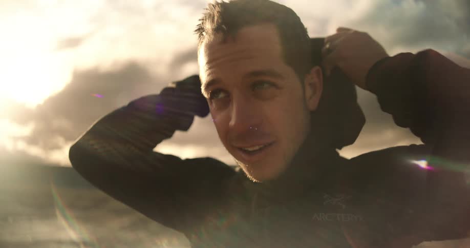 A closeup shows a man zipping his jacket and pulling the hood up with a lens flare appearing over him and sun in the background