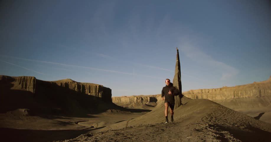 A person in running gear jogs across a barren hilly landscape under a clear blue sky with a tall thin rock formation in the background
