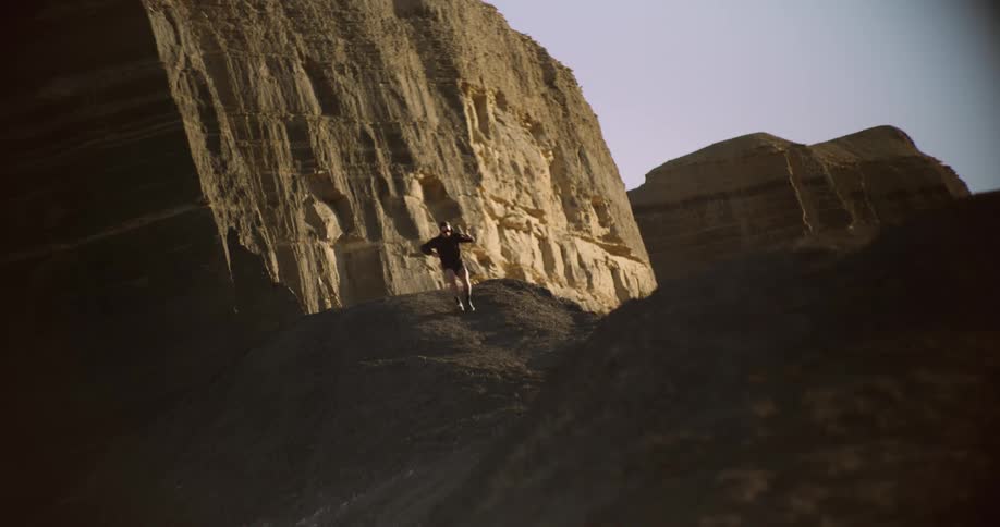 A person is running through a steep rocky hill with large rugged cliffs in the background illuminated by sunlight