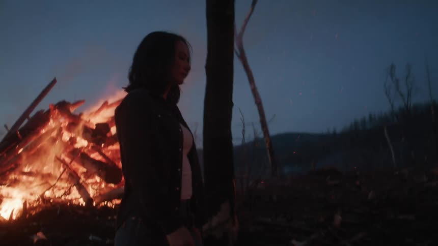 A silhouetted woman walks away from a blazing bonfire at night with trees visible in the darkness