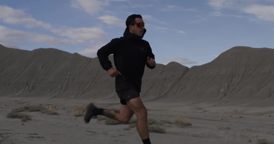 A person is running in a desert landscape with rugged hills in the background under a partly cloudy sky