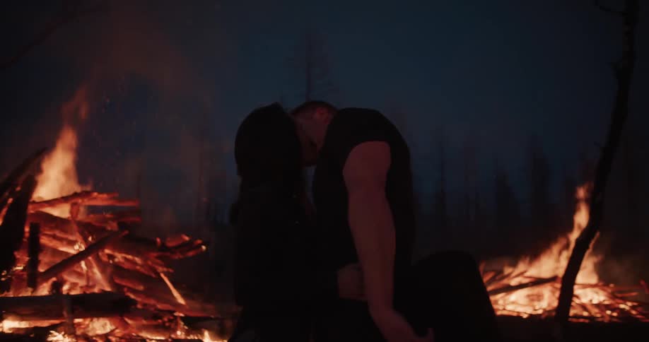 Man removing his cowboy hat to kiss woman in front of a large bonfire at night