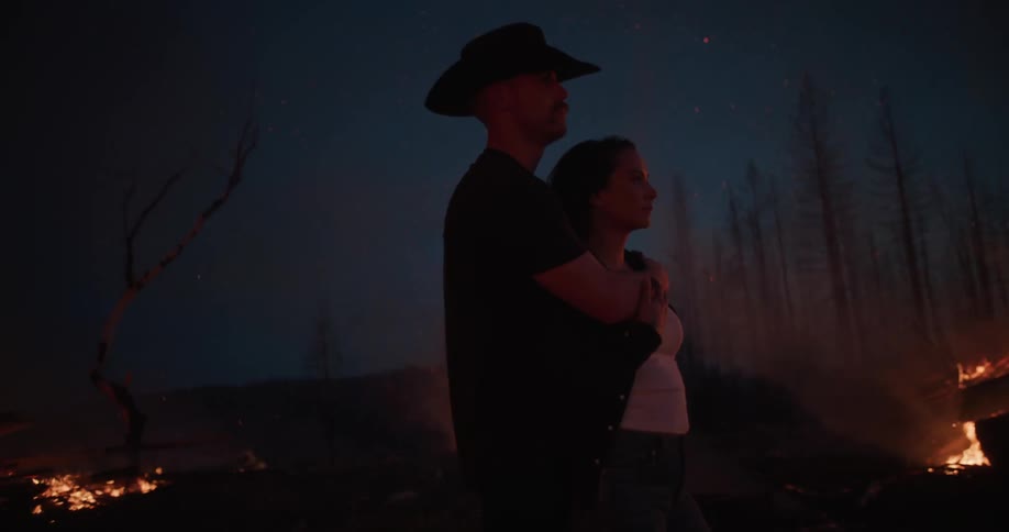 Man with hat and woman hugging in front of a fire in the background at night