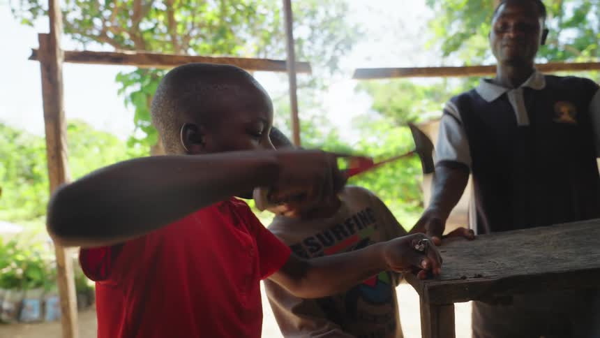 Boy with a hammer hammering a nail into a piece of wood accompanied by another child and an adult