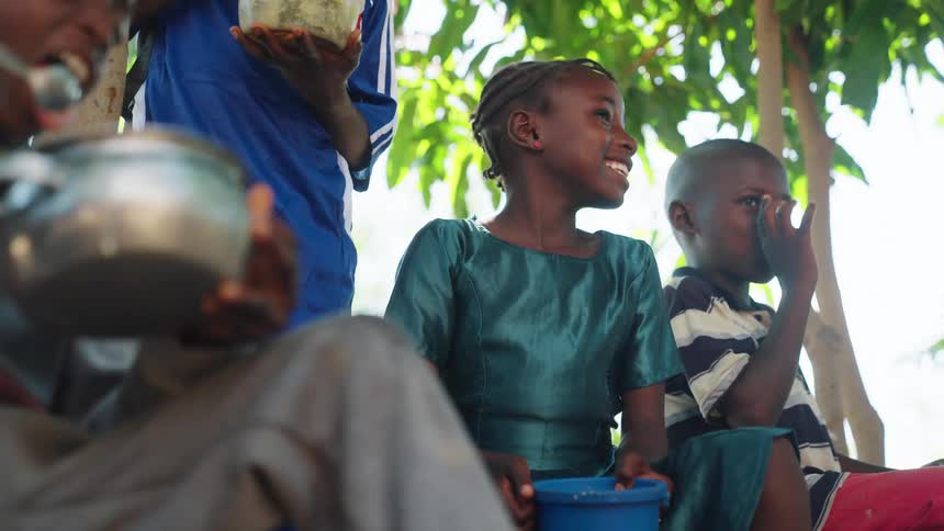 Children sit outdoors with one holding a blue container and another holding a metal pot surrounded by green foliage and wooden posts