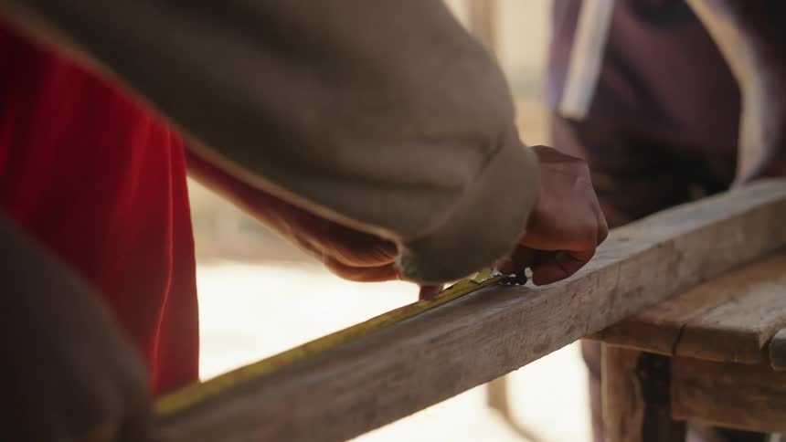 A mans hand is shown measuring a wooden plank on something being built and then cutting into it
