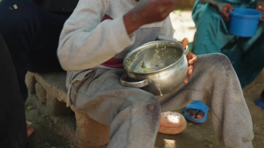 Boy sitting on a rock eating food from a pot with a spoon