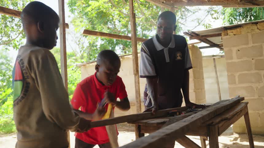 Two boys work with wood under the supervision of a man in an open sunny environment