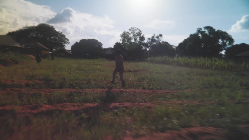 A child is running through a grassy field with trees and a few buildings in the background under a bright sunny sky