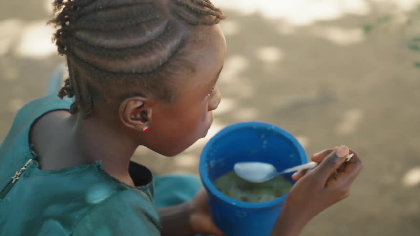A black girl is sitting outdoors and eating soup from a blue bowl