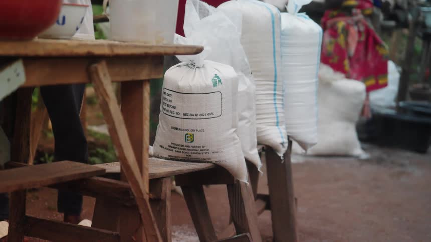 Several white sacks filled with grains labeled with text are placed on wooden stools in an outdoor setting