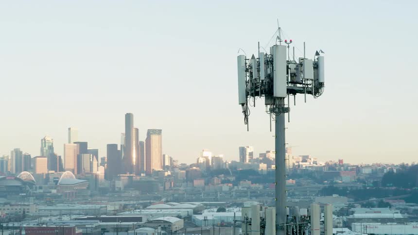 A telecommunications tower with multiple antennas stands in an urban area with a cityscape and downtown skyscrapers in the distance
