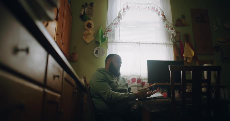 A man sits at a kitchen table flipping through papers and writing