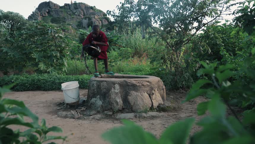 Woman standing edge of well pulling water from a bucket then transfering to another bucket