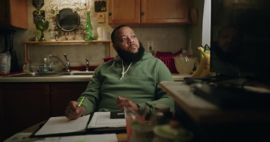 A person in a green hoodie sits at a kitchen table with a notebook pen and various papers appearing to work on taxes