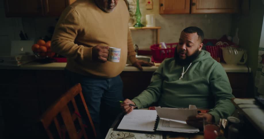 A man is sitting at a dining table in a cozy kitchen explaining paperwork to another person