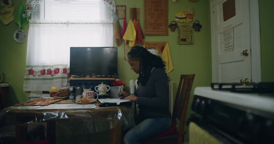 A woman is sitting at a dining table in a cozy kitchen working on paperwork with a pen