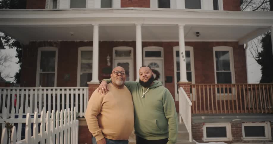 Father is embraced by his son standing in front of a house with a white picket fence