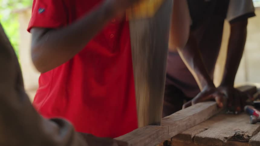 A boy using a saw to cut a piece of wood that is held diagonally by a person