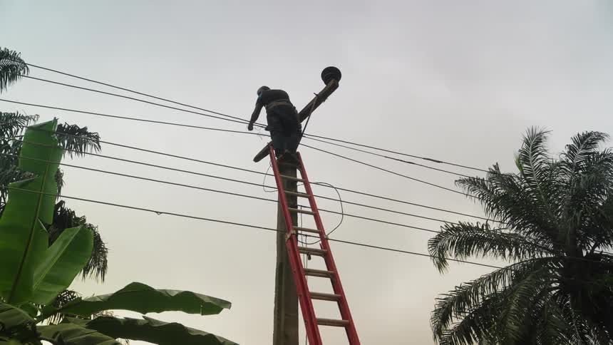 A worker on a red ladder leans against a utility pole adjusting equipment on the wires against a cloudy sky backdrop