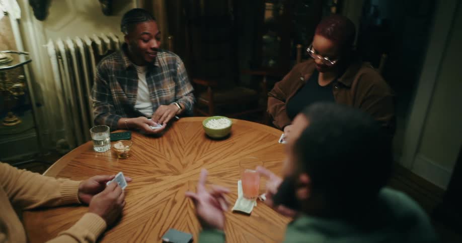 Three people are partially visible around a round table as they play cards with a fourth person in the background