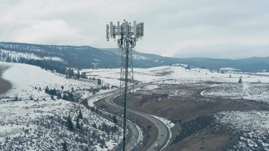 A cell tower stands prominently above snowy foothills as vehicles travel along a highway curving through the terrain below