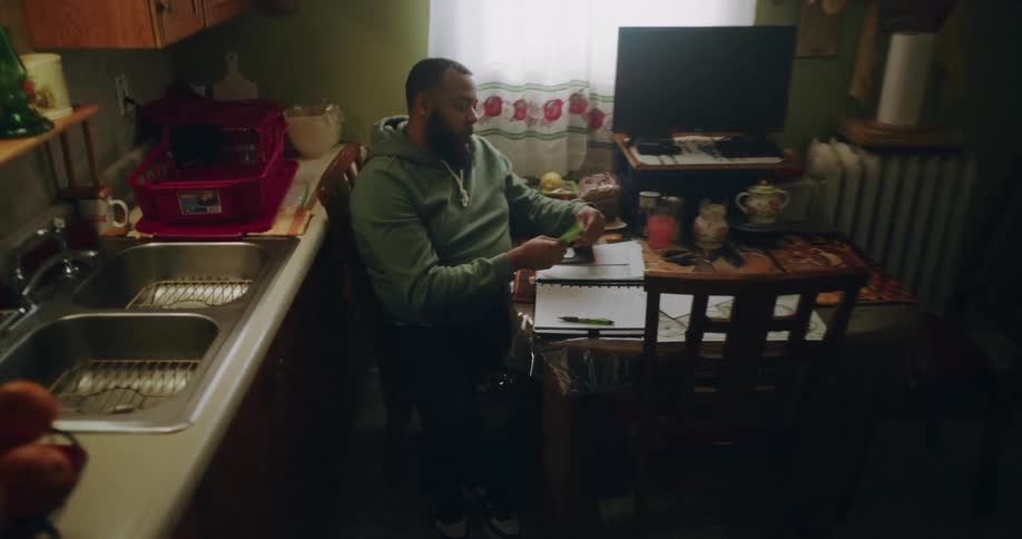 In a dimly lit kitchen a man in a green hoodie sits at a cluttered table reviewing papers and writing in a notebook