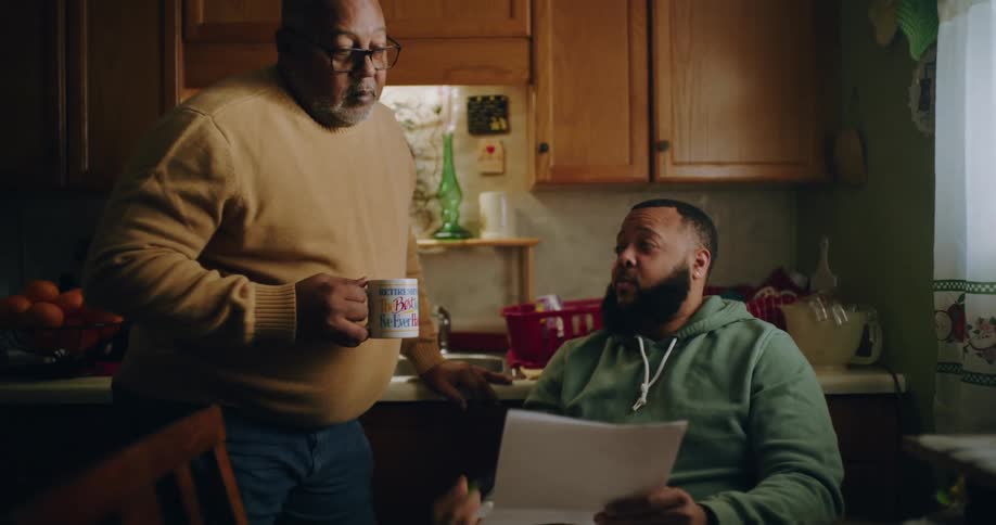 A man is sitting at a dining table in a cozy kitchen explaining paperwork to another person