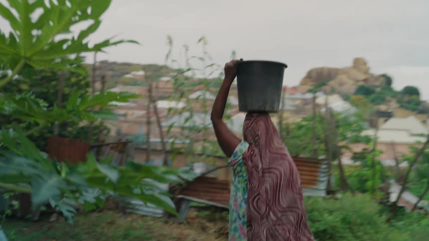 A woman walks through the yard balancing a bucket on her head